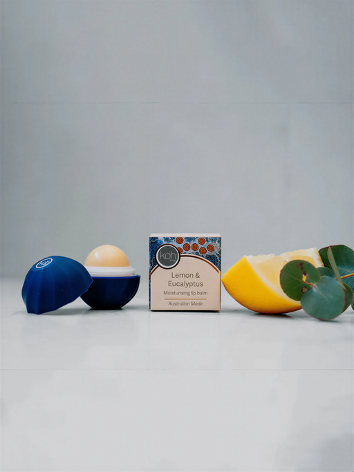 a box of lip balm, a blue container, a lemon, and a sprig of eucalyptus, all arranged on a white surface.