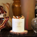 a white candle with text on it, placed on a wooden coaster, surrounded by various decorative items.