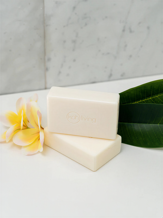 two rectangular bars of white soap with the word "Koh Living" printed on them, placed on a white surface next to a yellow flower and a green leaf.