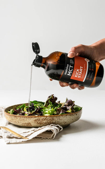 A hand is pouring a dark liquid, possibly a sauce or dressing, over a bowl of fresh salad greens.