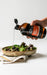A hand is pouring a dark liquid, possibly a sauce or dressing, over a bowl of fresh salad greens.