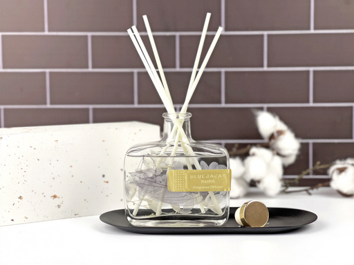 A clear glass bottle containing white reeds is placed on a black tray, with a white brick wall and some white cotton balls in the background.