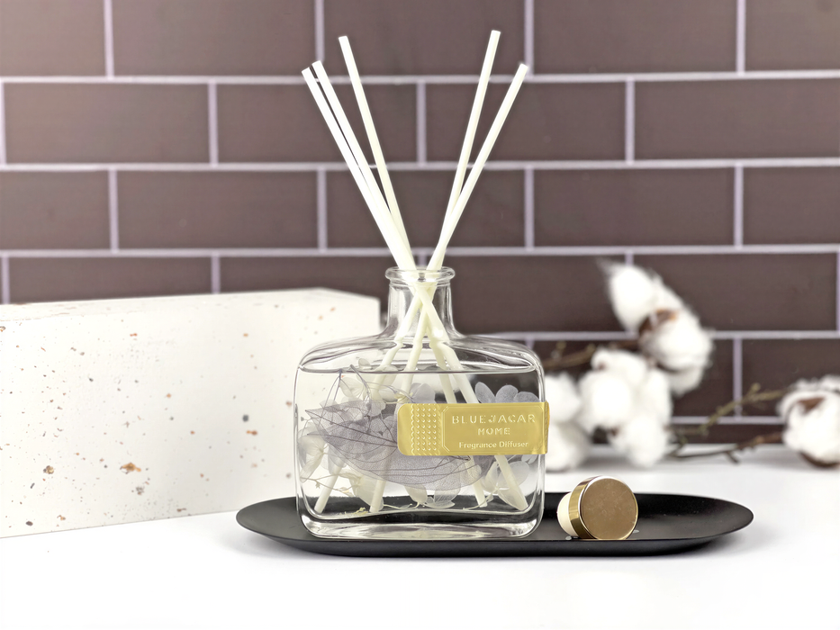 A clear glass bottle containing white reeds is placed on a black tray, with a white brick wall and some white cotton balls in the background.