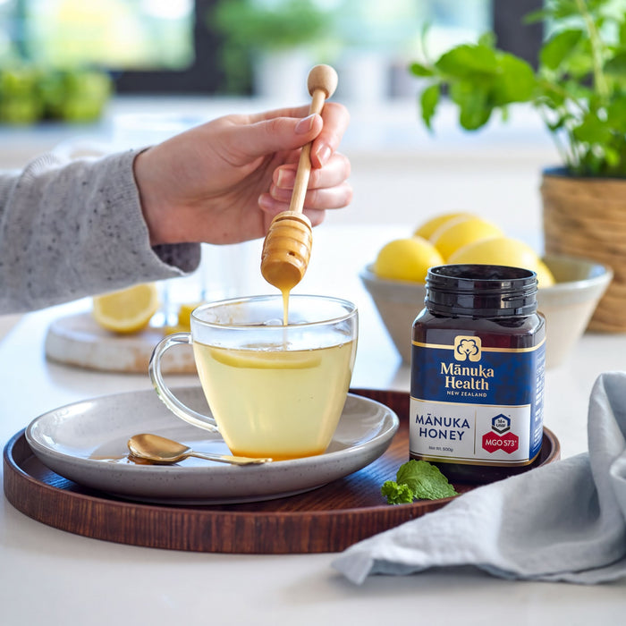A person's hand is pouring Manuka Honey into a glass of tea, with a spoon and a jar of Manuka Honey visible on the table.