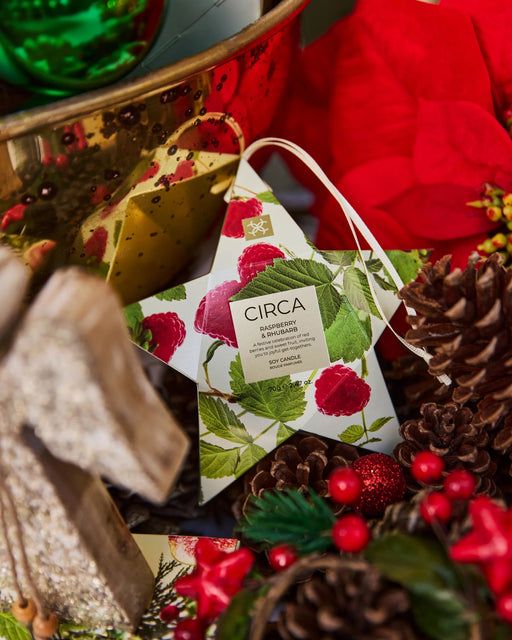 A star-shaped ornament with a label that reads "CIRCA RASPBERRY & RHUBSINI SINCE 1853" is surrounded by various holiday decorations, including pine cones, berries, and a green ornament.
