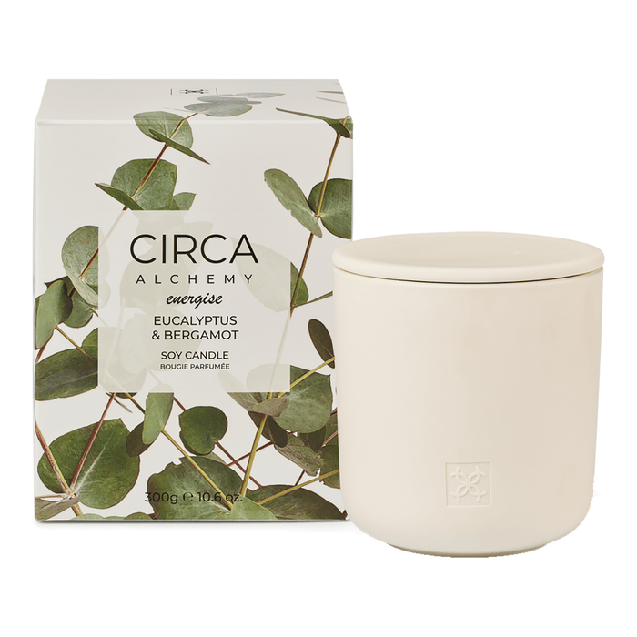 a white ceramic candle in a box with a green leaf design, and a white ceramic jar next to it.
