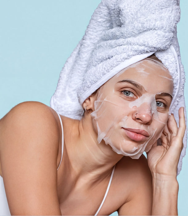 A woman with a white towel wrapped around her head and a sheet mask covering her face, looking directly at the camera with a serious expression.