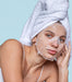 A woman with a white towel wrapped around her head and a sheet mask covering her face, looking directly at the camera with a serious expression.