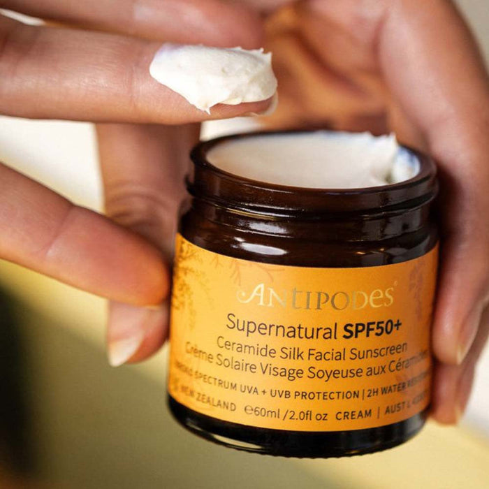 A person's hand is holding a jar of "Antipodes Supernatural SPF50+ Ceramide Silk Facial Sunscreen" cream.