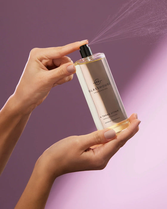 A person's hands holding a bottle of "A TAHA AFFAIR VANILLA CAFFEINE INTERIOR FRANCE" fragrance spray against a purple background.