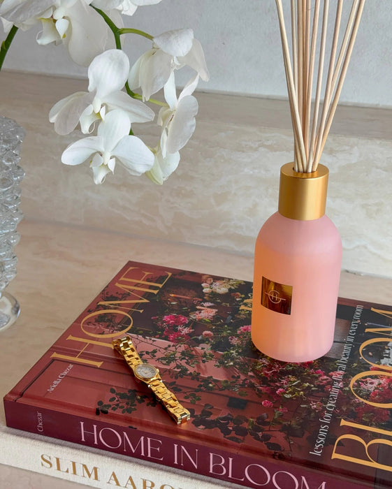 a pink diffuser with a gold-colored cap, a gold-colored watch, and a book titled "HOME IN BLOOM" on a table.