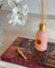 a pink diffuser with a gold-colored cap, a gold-colored watch, and a book titled "HOME IN BLOOM" on a table.