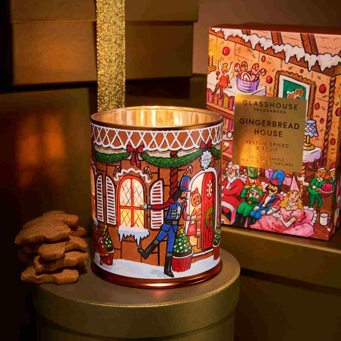 A decorative gingerbread house candle in a tin, with a stack of gingerbread cookies next to it, and a box of the same candle in the background.
