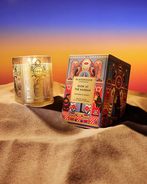 a glass candle holder and a box of Glasshouse Dusk at the Kasbah scented candle, both placed on a sandy surface against a colorful sunset sky.