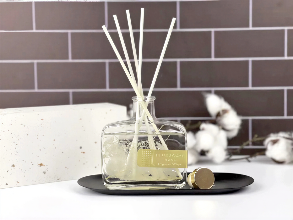 A glass diffuser with white reeds and a label that reads "AUBURN HOME Fragrance Diffuser" is placed on a black tray against a brick wall background.