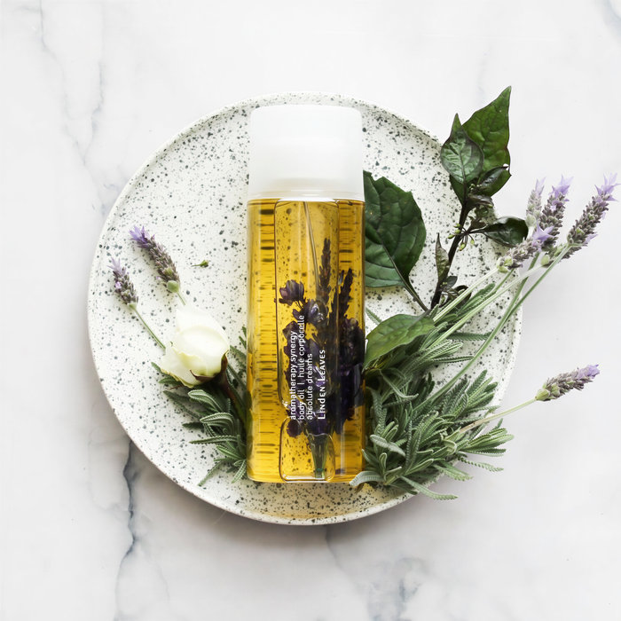 A round white plate with a spiky texture holds a clear bottle containing a yellow liquid surrounded by purple flowers and green leaves.