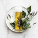 A round white plate with a spiky texture holds a clear bottle containing a yellow liquid surrounded by purple flowers and green leaves.