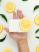 A hand holding a bar of soap with a slice of lemon on top, surrounded by lemon slices and green leaves.