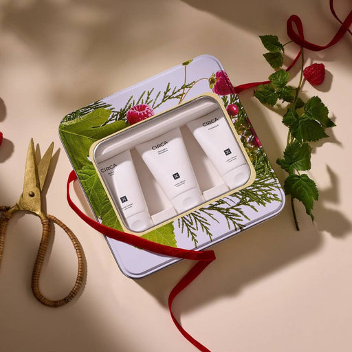 A set of three white tubes of CICA skincare products, each with a black label, arranged in a decorative tin box with a red ribbon. The tin box is placed on a white surface, with a pair of scissors and some greenery visible in the background.