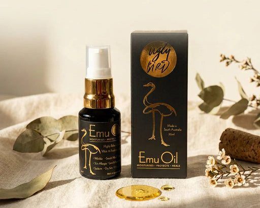 a bottle of Emu Oil and its packaging, both placed on a beige surface with some leaves and flowers in the background.
