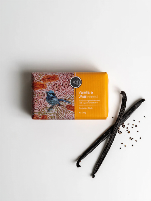 a bar of soap with a bird design, along with a pair of black vanilla pods and some seeds on a white background.