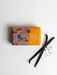 a bar of soap with a bird design, along with a pair of black vanilla pods and some seeds on a white background.