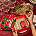 A person's hands holding a gift box with a red and green design, surrounded by a variety of wine glasses, a plate of food, and jewelry on a table.