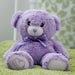 A purple teddy bear with a heart-shaped tag sits on a green surface, with a blurred green and white patterned background.