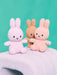 Three plush toy rabbits in different colors - pink, brown, and white - sitting on a white surface against a teal background.