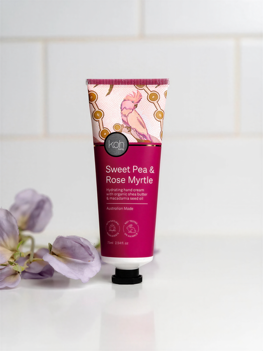 A tube of Sweet Pea & Rose Myrtle hand cream is displayed on a white countertop, with purple flowers in the background.