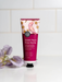 A tube of Sweet Pea & Rose Myrtle hand cream is displayed on a white countertop, with purple flowers in the background.