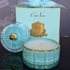 a turquoise glass candle holder with a white candle inside, placed next to a box with a gold label and a blue ribbon.