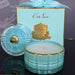 a turquoise glass candle holder with a white candle inside, placed next to a box with a gold label and a blue ribbon.
