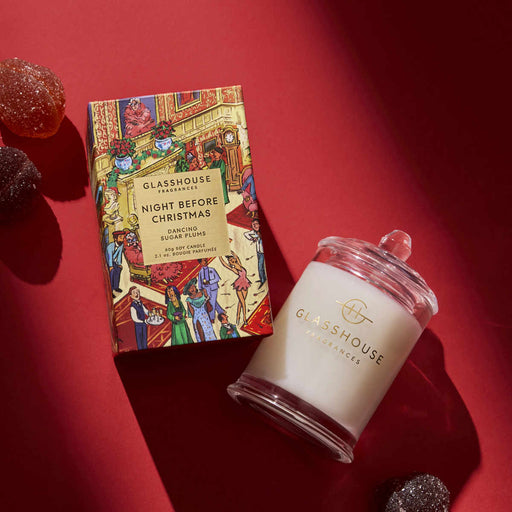 a box of Glashouse Fragrances Night Before Christmas Sugar Plums and a candle in a glass jar, both placed on a red surface.