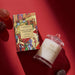 a box of Glashouse Fragrances Night Before Christmas Sugar Plums and a candle in a glass jar, both placed on a red surface.