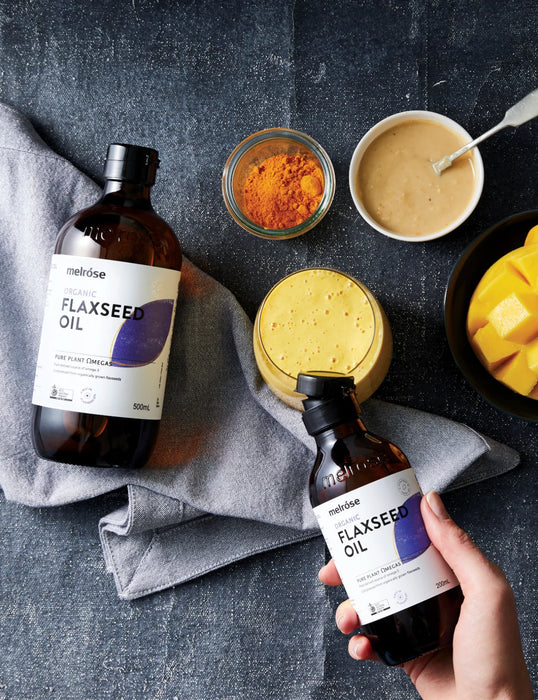 a hand holding two bottles of melrose FLAXSEED OIL, surrounded by various ingredients and a bowl of sauce on a dark surface.