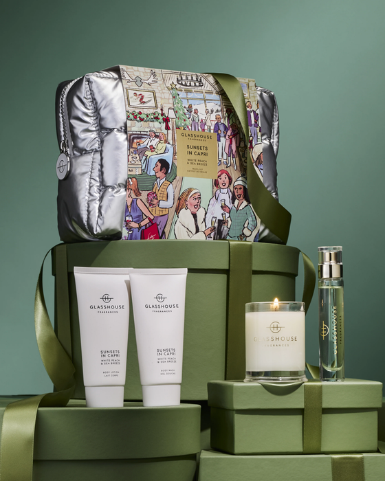 A gift set with a metallic bag featuring a colorful illustration, two white bottles of GLASSHOUSE fragrances, a candle, and a bottle of GLASSHOUSE perfume.