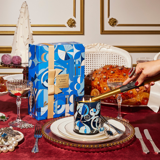 a table set for a formal dinner, with a candle, wine glasses, and a large cake. A hand is holding a lighter, preparing to light the candle. The table is adorned with a blue and white patterned box, and the background includes a white wall with gold trim.