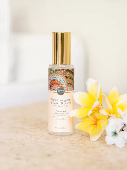 A bottle of "Native Frangipani & Peach Blossom" perfume with a gold cap, placed on a table next to a bunch of yellow flowers.
