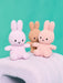 Three plush toy rabbits in different colors - pink, brown, and white - sitting on a white surface against a teal background.