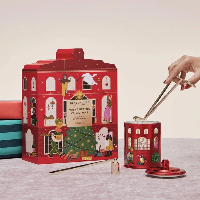 a red box with a Christmas-themed illustration, a hand holding a pair of tongs, and a red tin can with a lid, all placed on a table.