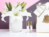 a bouquet of white lilies in a glass vase, along with two small perfume bottles, all placed on a white surface.