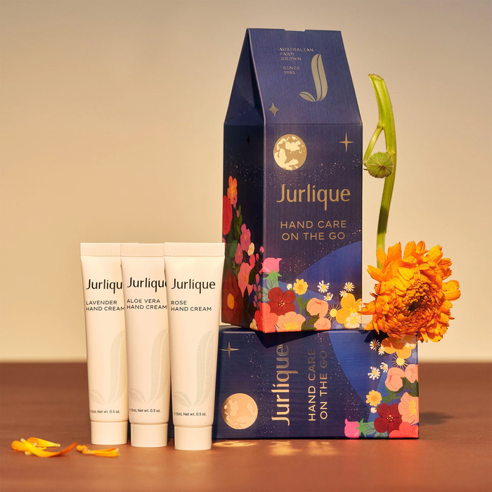 a collection of beauty products, including three tubes of Jurlique hand cream and a box of hand care on the go. The products are arranged on a wooden surface, with a single orange flower adding a pop of color to the scene.