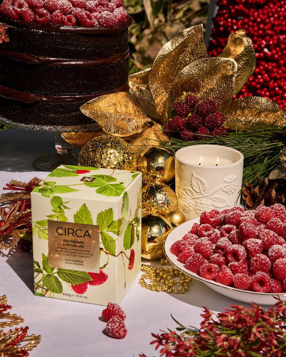 a table with a variety of items, including a box of CIRCA raspberry-flavored tea, a bowl of raspberries, a candle, and a cake. The table is decorated with gold and red ornaments, and the overall scene appears to be a festive or holiday-themed setting.