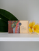 A bar of soap with a colorful, abstract design sits on a white surface, accompanied by a yellow flower and a green leaf.