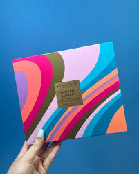 A hand holding a colorful box with a gold label that reads "Fragrance Library" against a blue background.