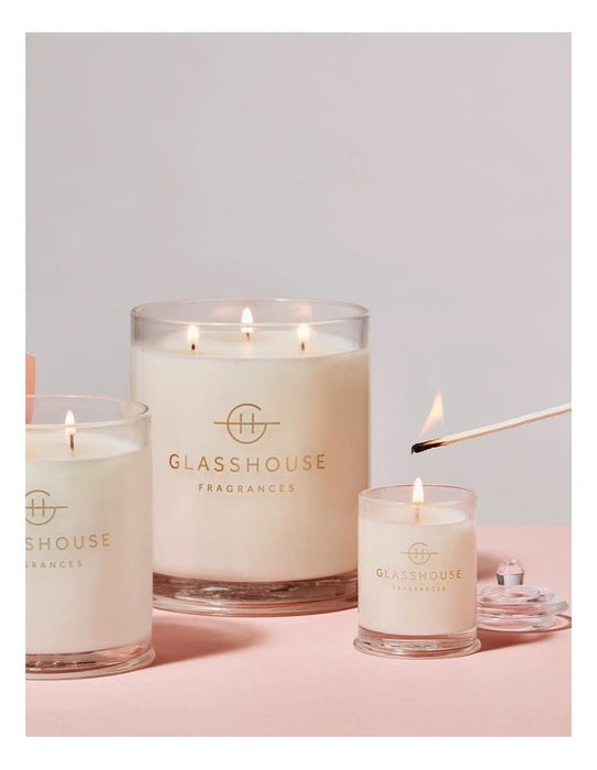 Three large glass jars with lit candles, each labeled with the "GLASSHOUSE Fragrances" logo, placed on a pink background.