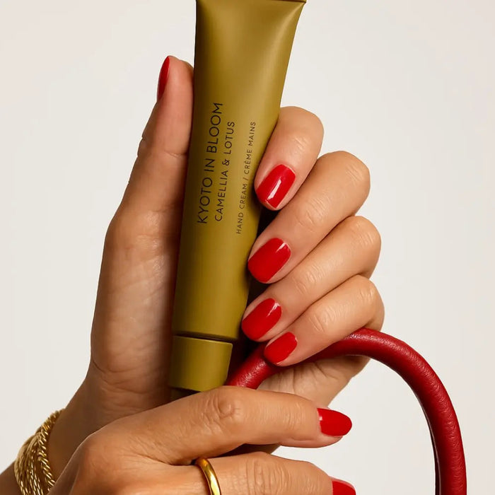 A person's hand holding a tube of lotion with red nail polish, and a red purse or bag is visible in the background.
