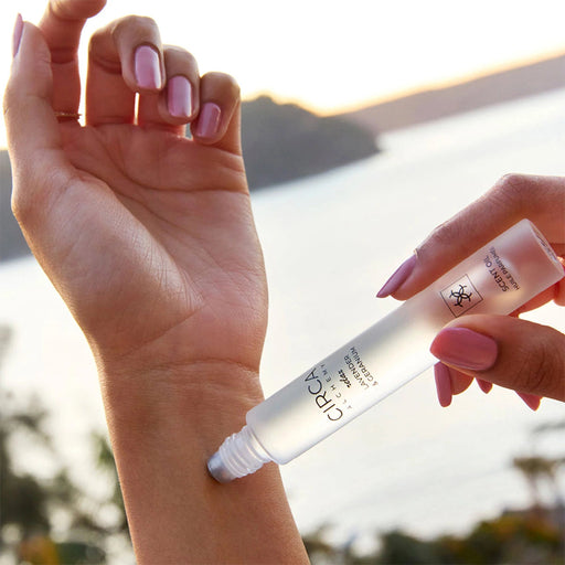 A person's hand applying a product to their wrist, with a body of water and a sunset in the background.