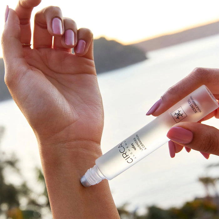A person's hand applying a product to their wrist, with a body of water and a sunset in the background.